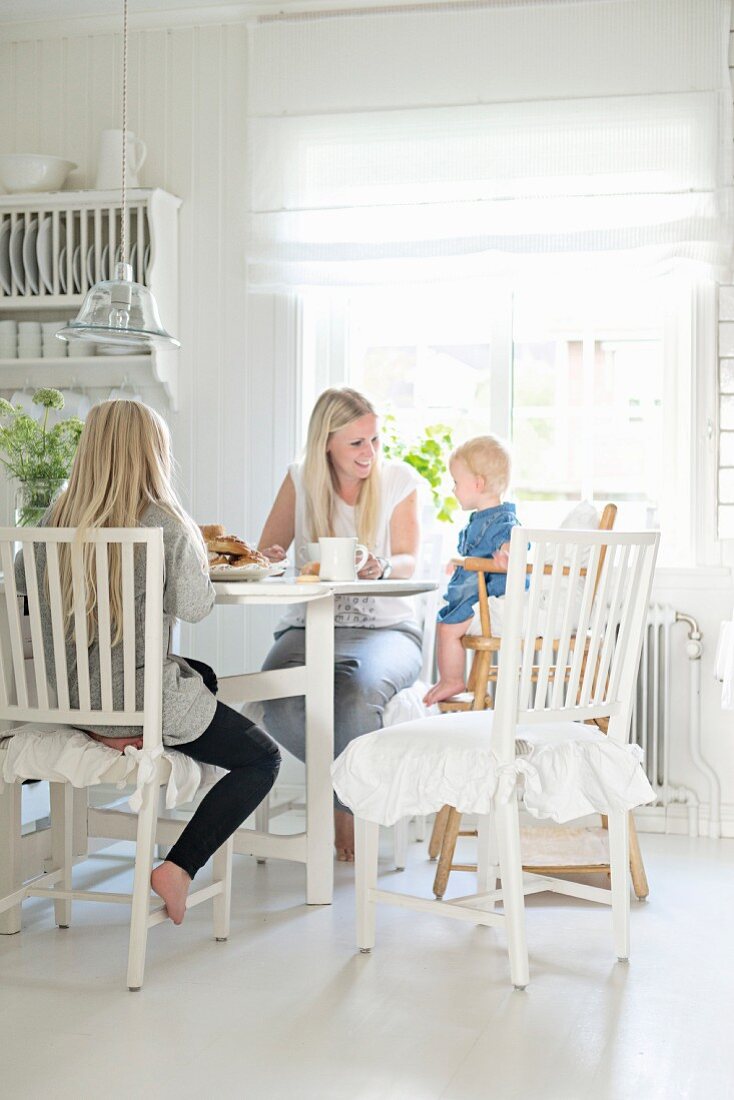 Mutter mit Tochter und Kleinkind im Kinderstuhl an Esstisch in weisser Landhausküche