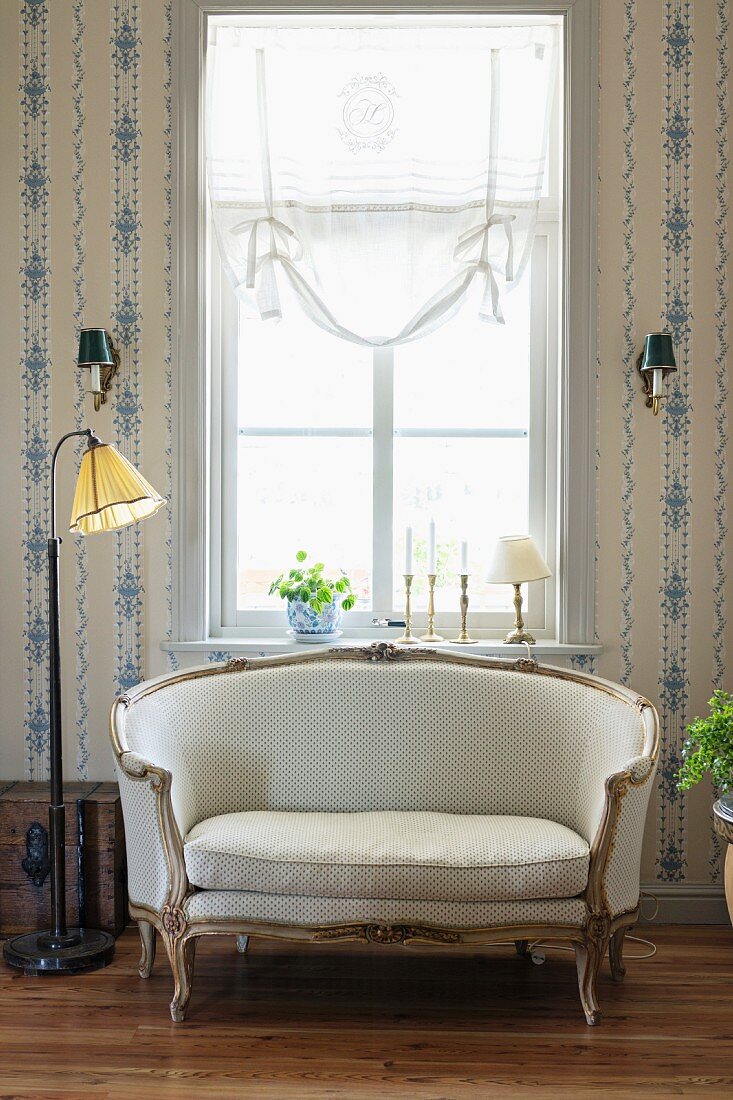 Rococo sofa and simple standard lamp in front of window flanked by wallpapered walls