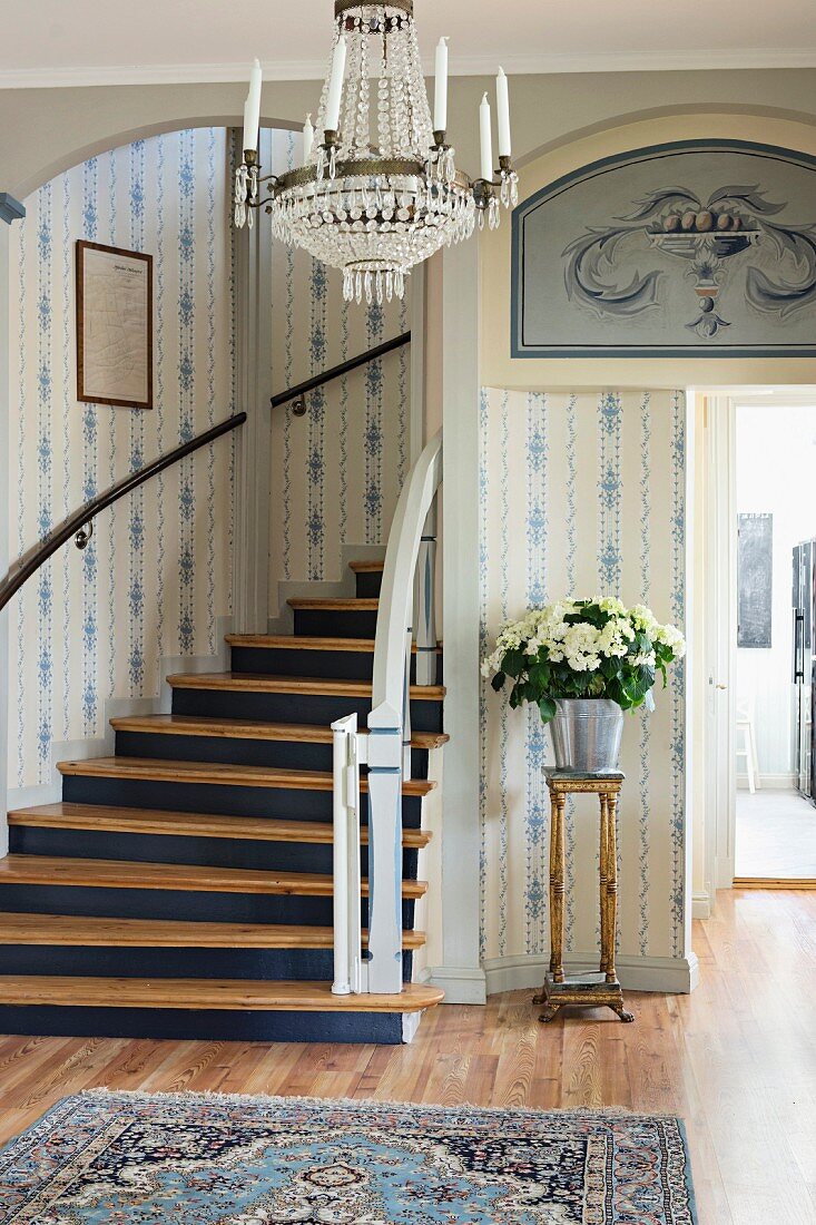 Eingangsbereich mit weiss-blauer Tapete, geschwungener Treppe und Blumenstrauss in silberner Vase auf Blumenständer