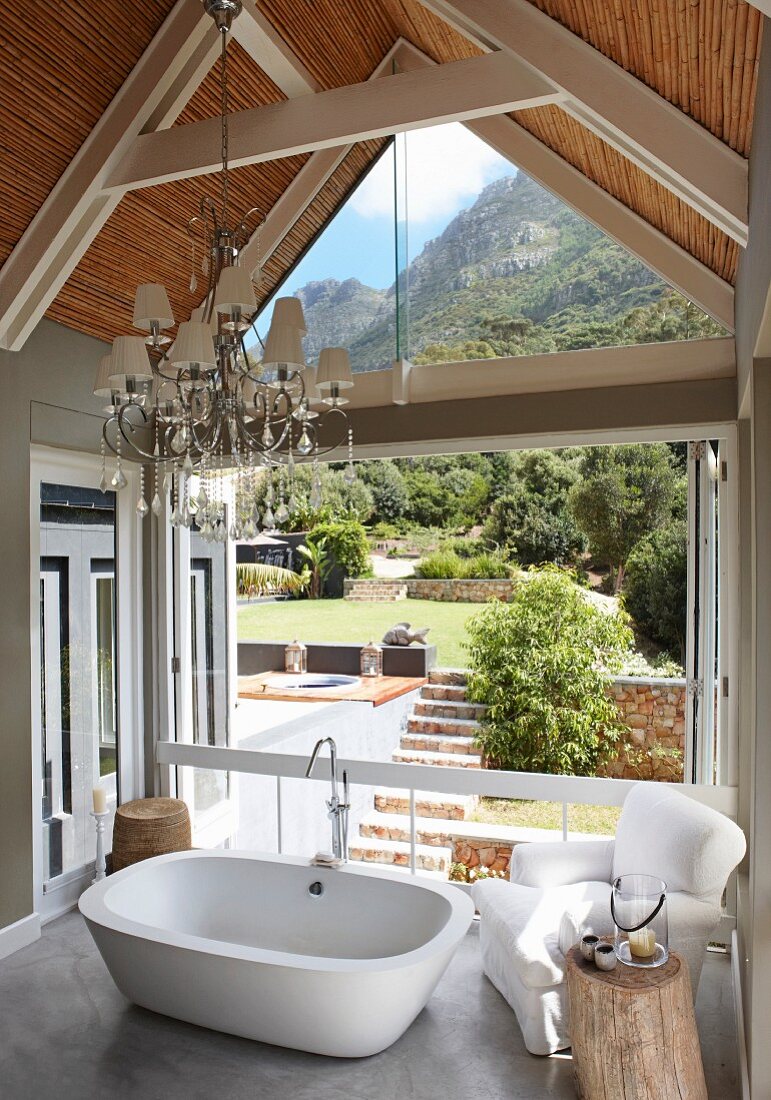 Free-standing bathtub and upholstered armchair on gallery in front of open folding doors with view into sunny garden