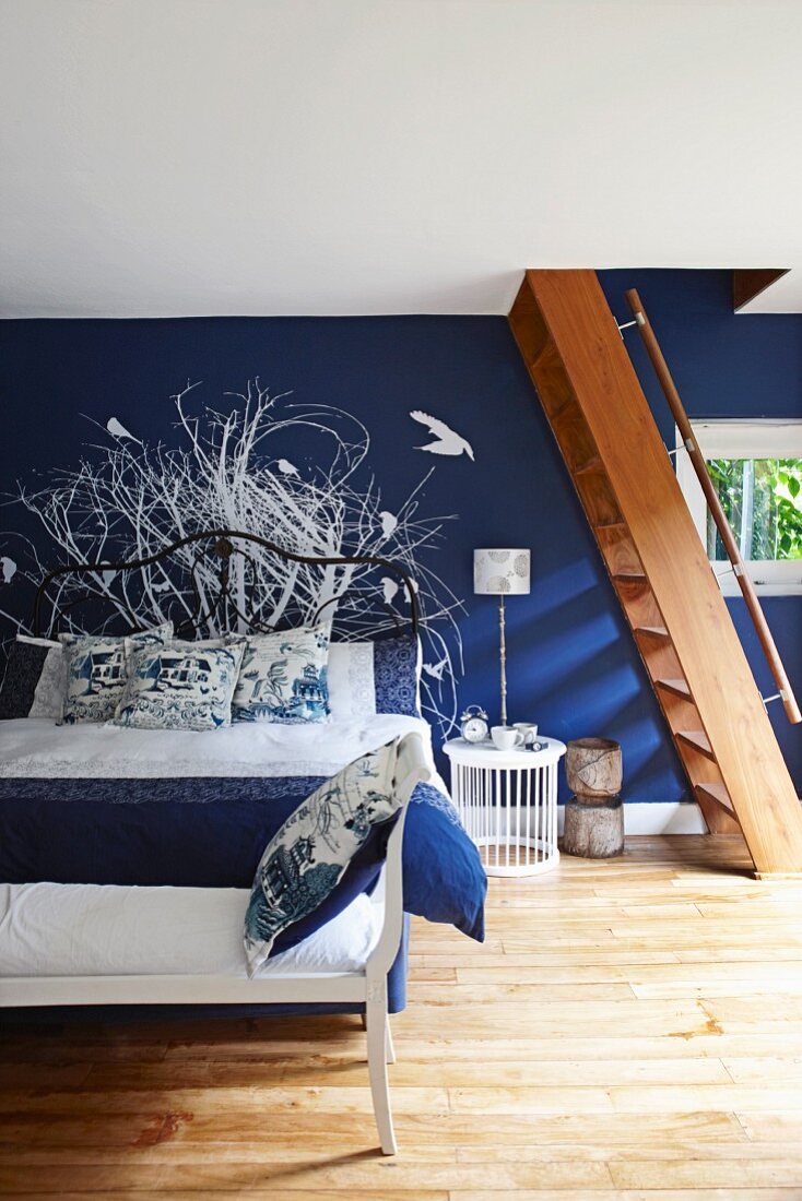 Schlafzimmer mit dunkelblauer Wand und weißem Wandgemälde, davor Holztreppe, im Vordergrund teilweise sichtbare Bank vor Doppelbett