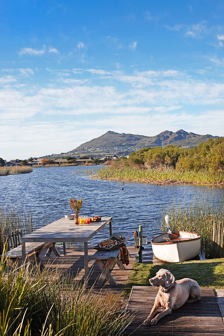 Dog in rustic seating area on wooden deck next to idyllic lake with rowing boat on shore and view of mountains