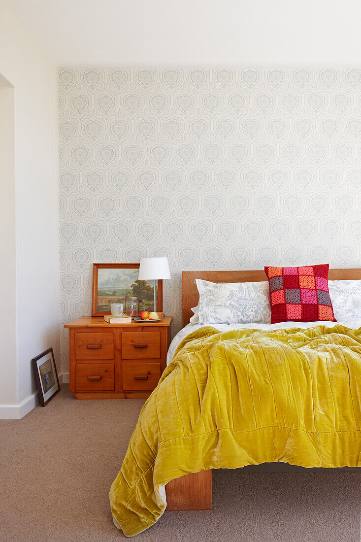 Double bed with yellow bedspread and small chest of drawers against wall with ornamental, patterned wallpaper