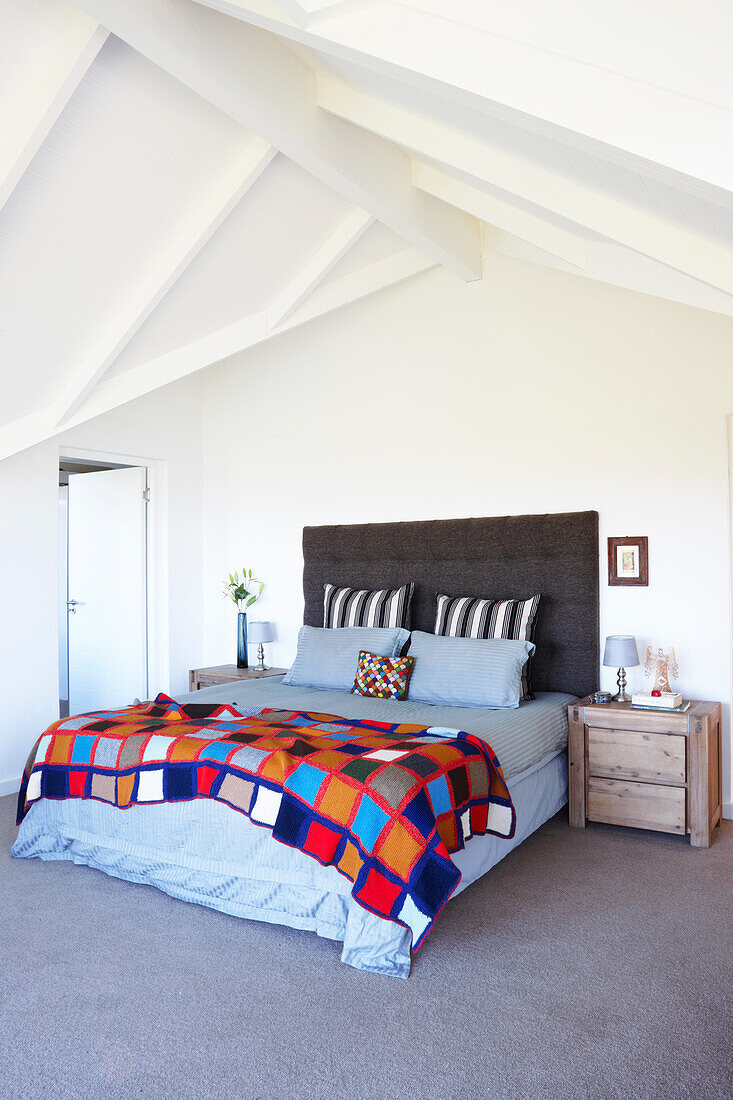 Double bed with button-tufted headboard and colourful patchwork bedspread below white-painted ridge beam