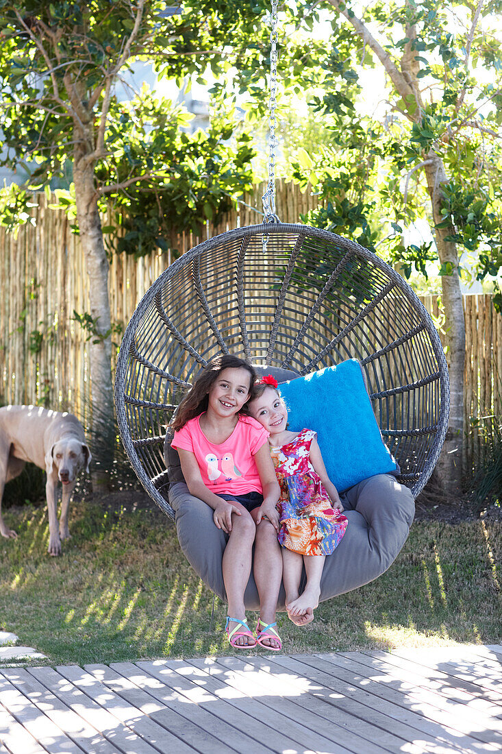 Two smiling girls sitting in wicker hanging chair in summer garden with dog in background