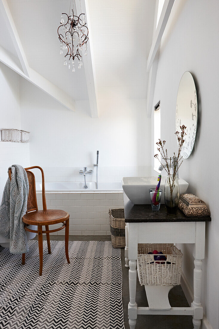 Rustic washstand with countertop sink and Thonet chair in front of fitted bathtub in white, attic bathroom