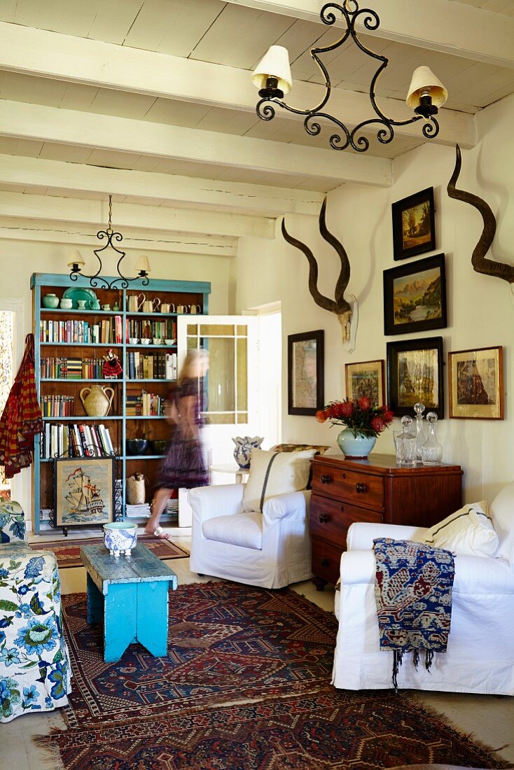 Sessel mit weisser Husse, dazwischen Kommode vor Wand unter gerahmten Bildern und Tiertrophäe, in ländlichem Wohnzimmer, mit weiss gestrichener Holzdecke