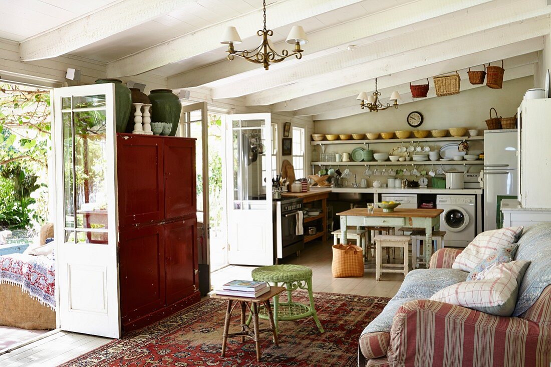 Ländlicher Wohnraum, mit weissen Holzbalkendecke, Polstercouch mit Kissen gegenüber offenen Terrassentüren, im Hintergrund Küche