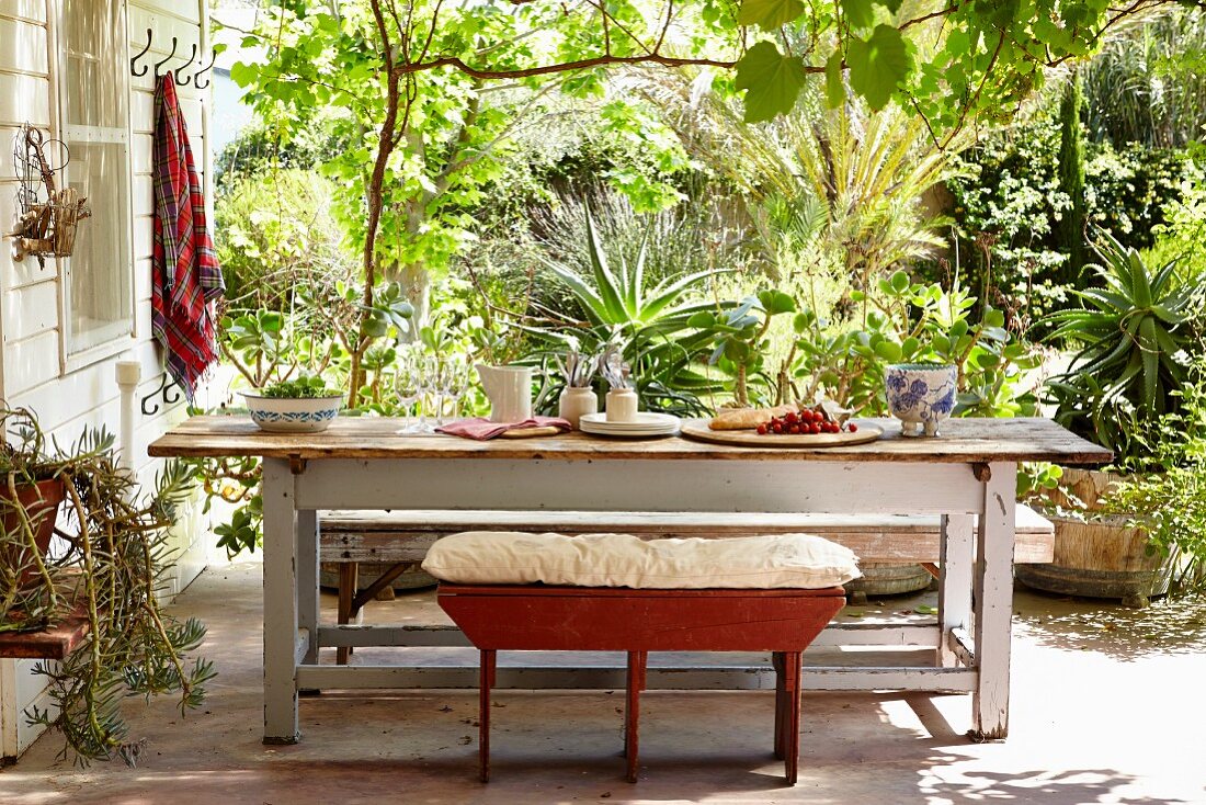 Bank mit Sitzkissen vor rustikalem Tisch auf Terrasse, im Hintergrund tropische Pflanzen in sonnigem Garten