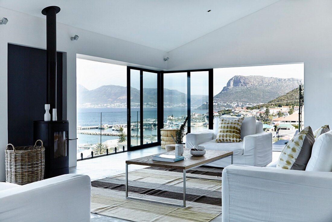 Elegant living room with white sofa set, delicate coffee table, partially glazed facade with sliding doors and panoramic view of mountainous coast