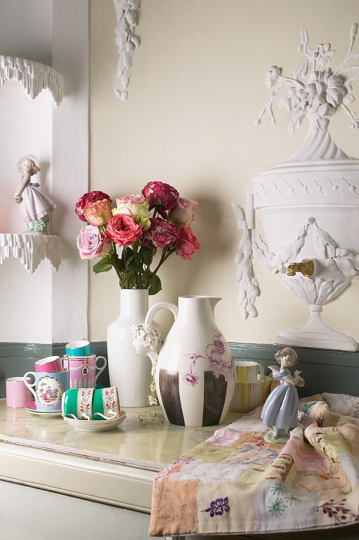 Collection of delicately patterned china teacups, china figurines and vases with bouquet of roses on marble cabinet top against wall with stucco reliefs