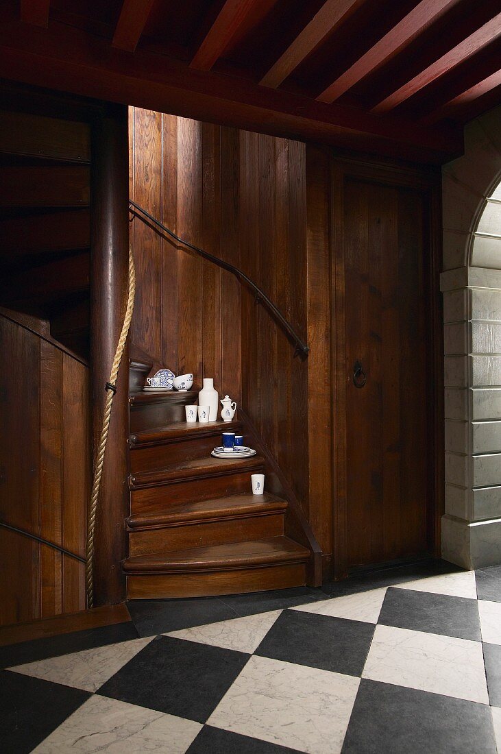 Porzellangeschirr auf den Stufen einer eingebauten Wendeltreppe aus dunklem Holz, davor ein Gang mit schwarzweissem Schachbrettboden