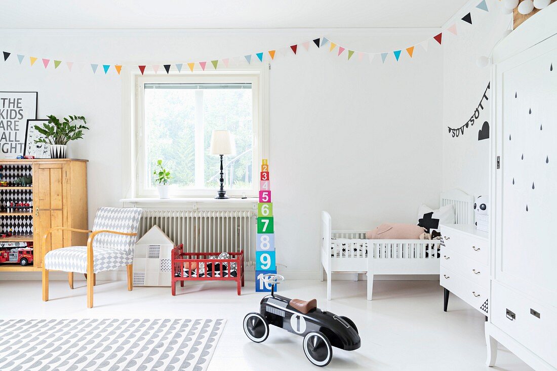 Grosszügiges Kinderzimmer mit bunter Wimpelgirlande und Retro Spielzeugauto, im Hintergrund rotes Puppenbett und weißes Gitterbett