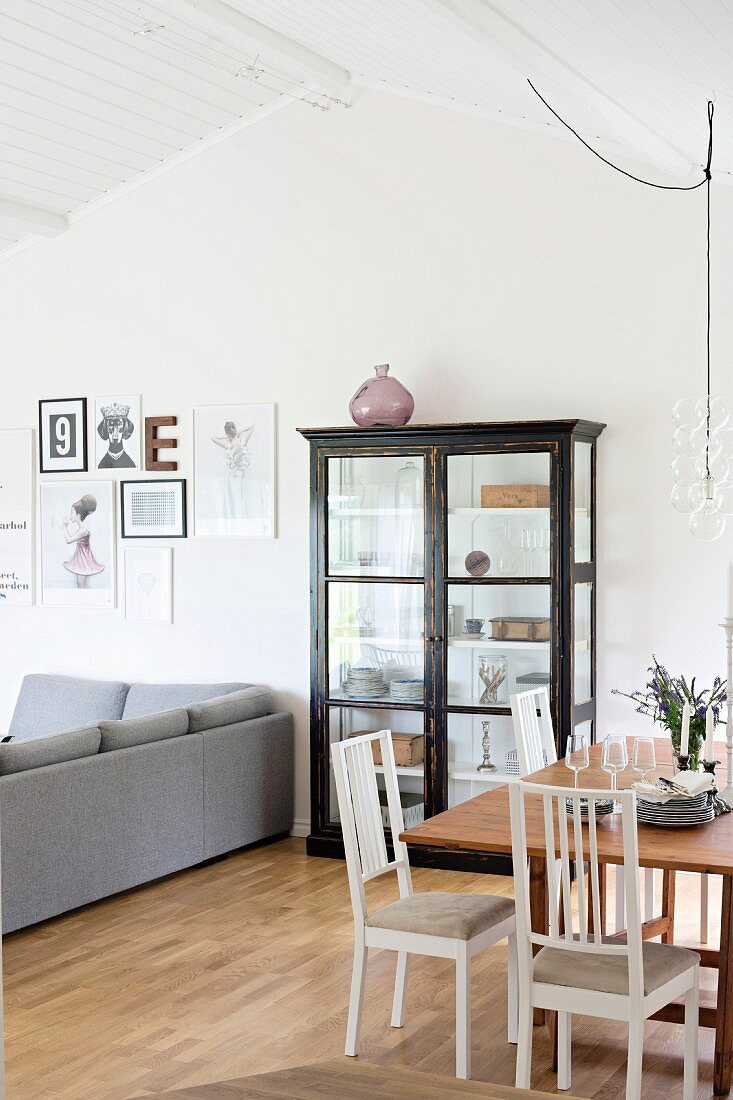Essplatz mit weissen Stühlen um Holztisch und Vitrinenschrank neben grauem Polstersofa in offenem Wohnraum