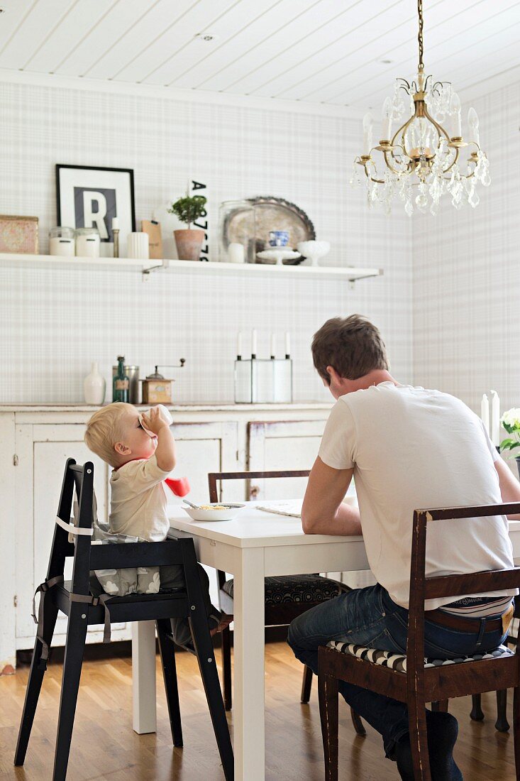 Father and child in high chair at white dining table below crystal chandelier