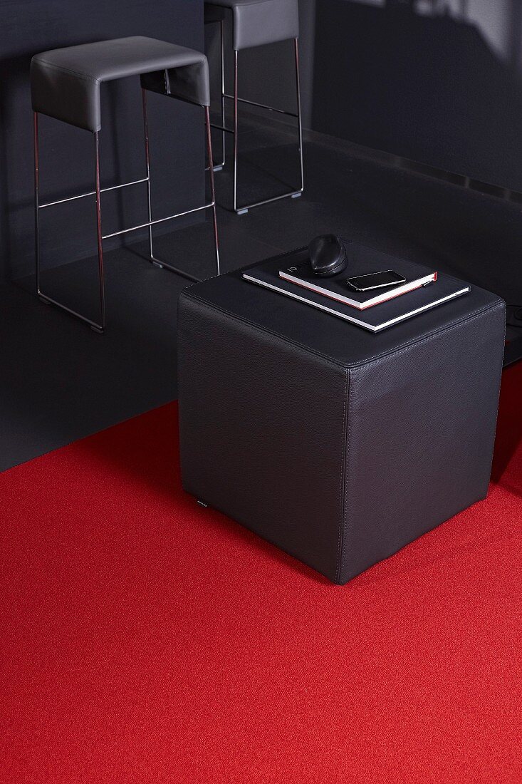 Grauer Designer-Sitzwürfel auf rotem Teppich und Barhocker im Hintergrund