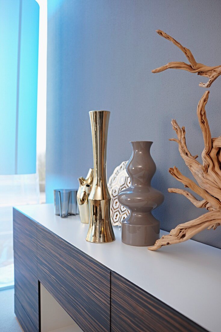 Elegante Vasen verschiedener Stilrichtungen und dekorative Wurzel auf Sideboard mit Edelholzfront