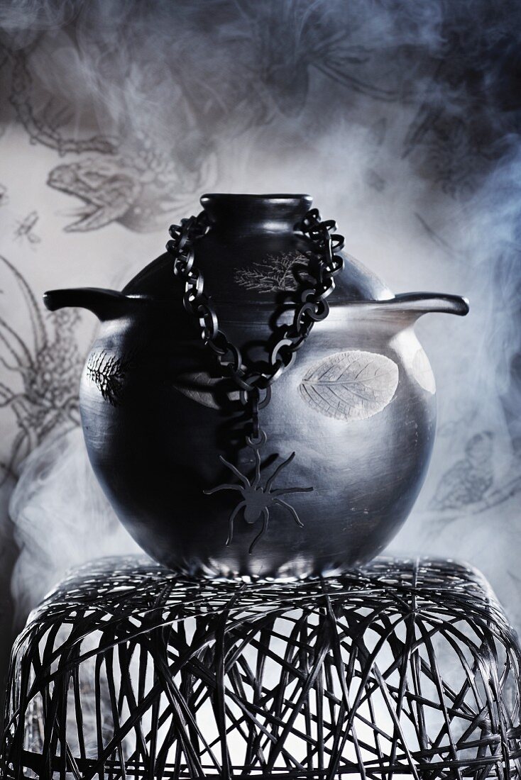 Düstere Inszenierung eines schwarzen Keramiktopfes, Kette mit Spinnenfigur und Hocker aus Metallgeflecht vor Rauchschwaden