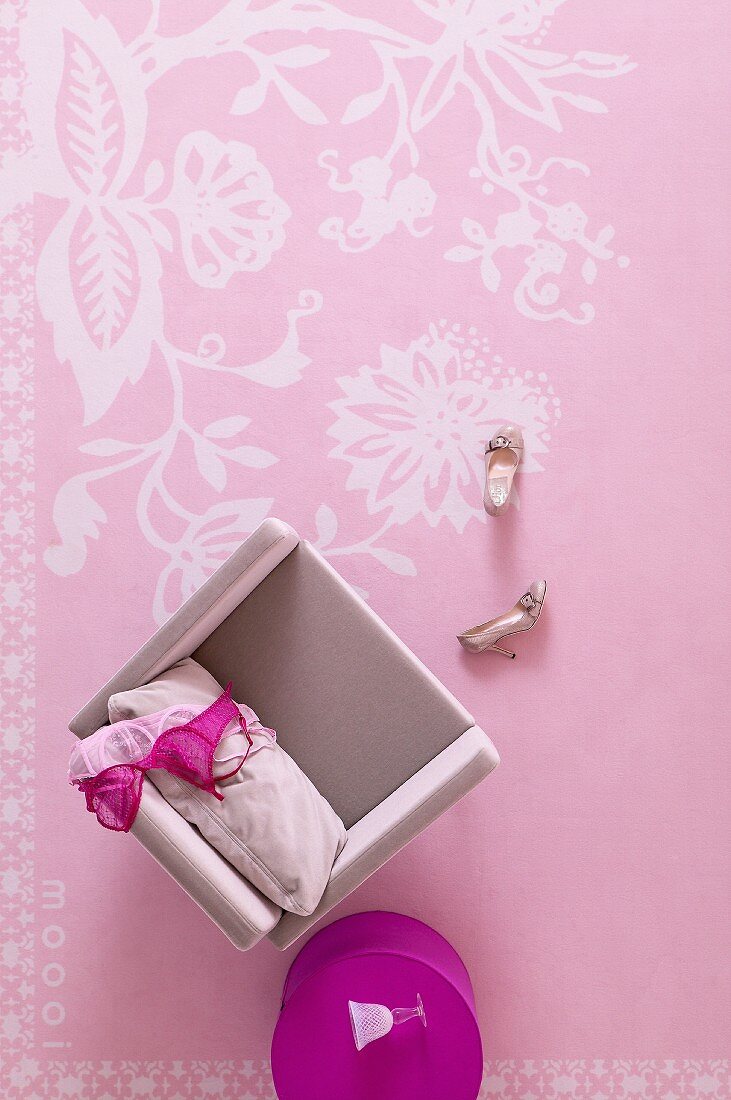 Blick von oben auf rosa Sessel mit Damenwäsche und fuchsiafarbener Beistelltisch auf rosa Teppich mit weißem, floralem Muster