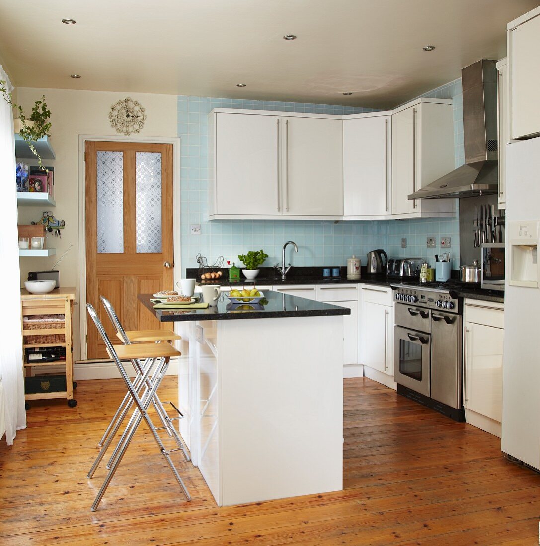 Moderne, weiße Einbauküche mit schwarzen Arbeitsflächen und hellblau gefliester Rückwand; mittig der Frühstückstresen