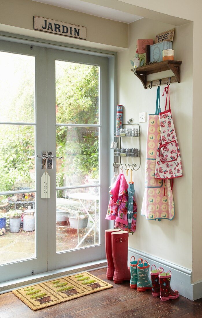 Kindergarderobe mit bunten Schürzen und Gummistiefeln neben Doppelfenstertür zum Garten mit französischem Vintage Schild