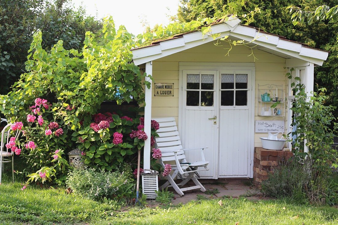 Romantic, climber-covered, wooden summer house painted pastel yellow with armchair under porch