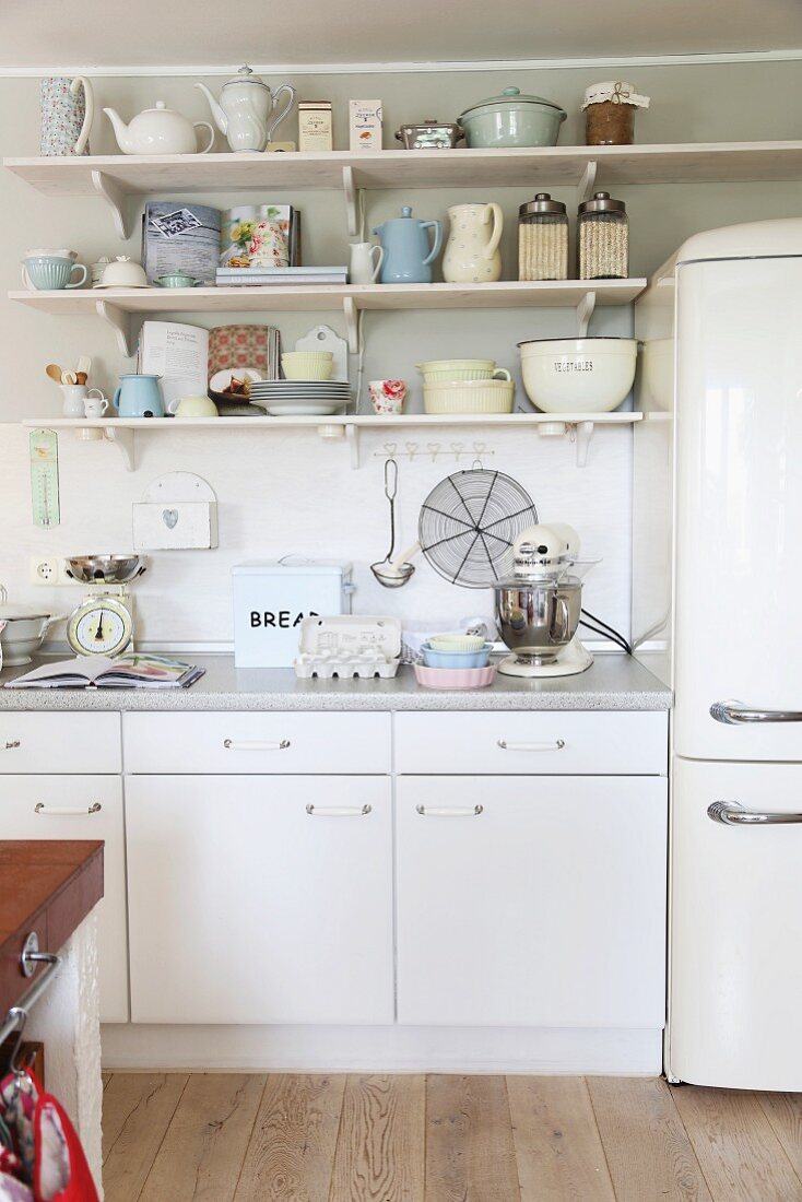 Simple, white kitchen counter with vintage crockery on wall-mounted shelves and retro-style fridge