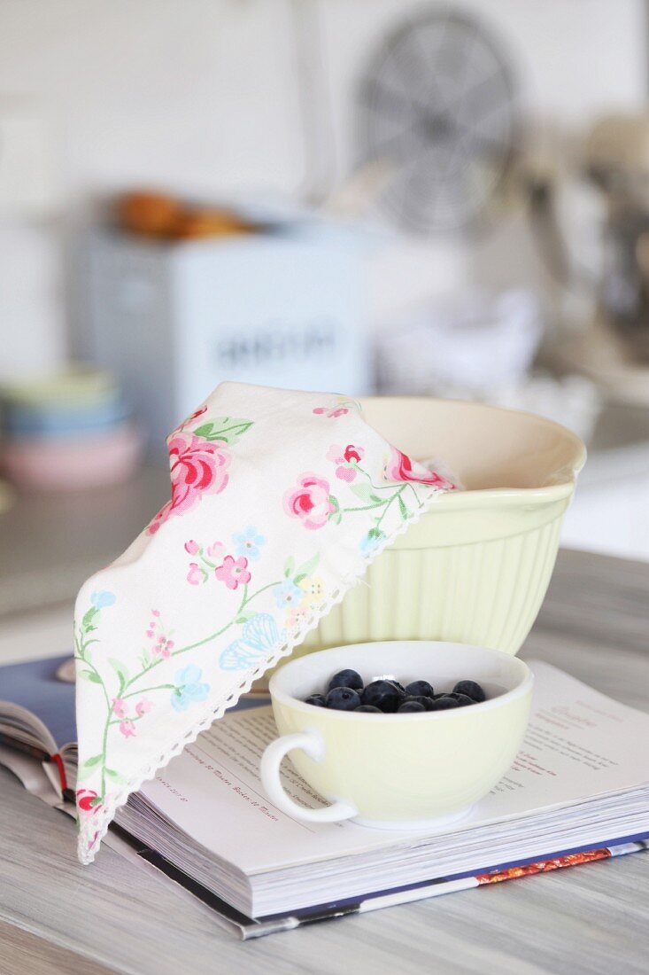 Tasse mit frischen Blaubeeren und Tuch mit Blumenmuster in Teigschüssel auf Rezeptebuch