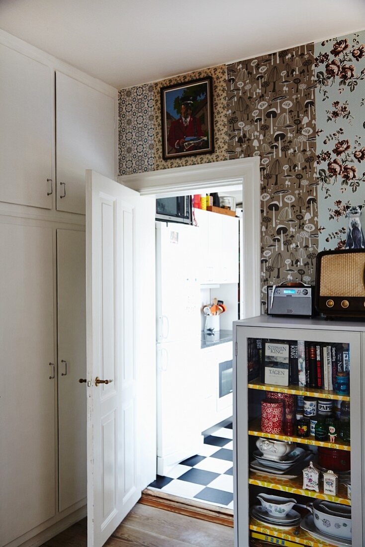 Vitrinenschrank vor Wand mit verschieden gemusterten Tapetenbahnen, seitlich offene Tür und Blick in die Küche