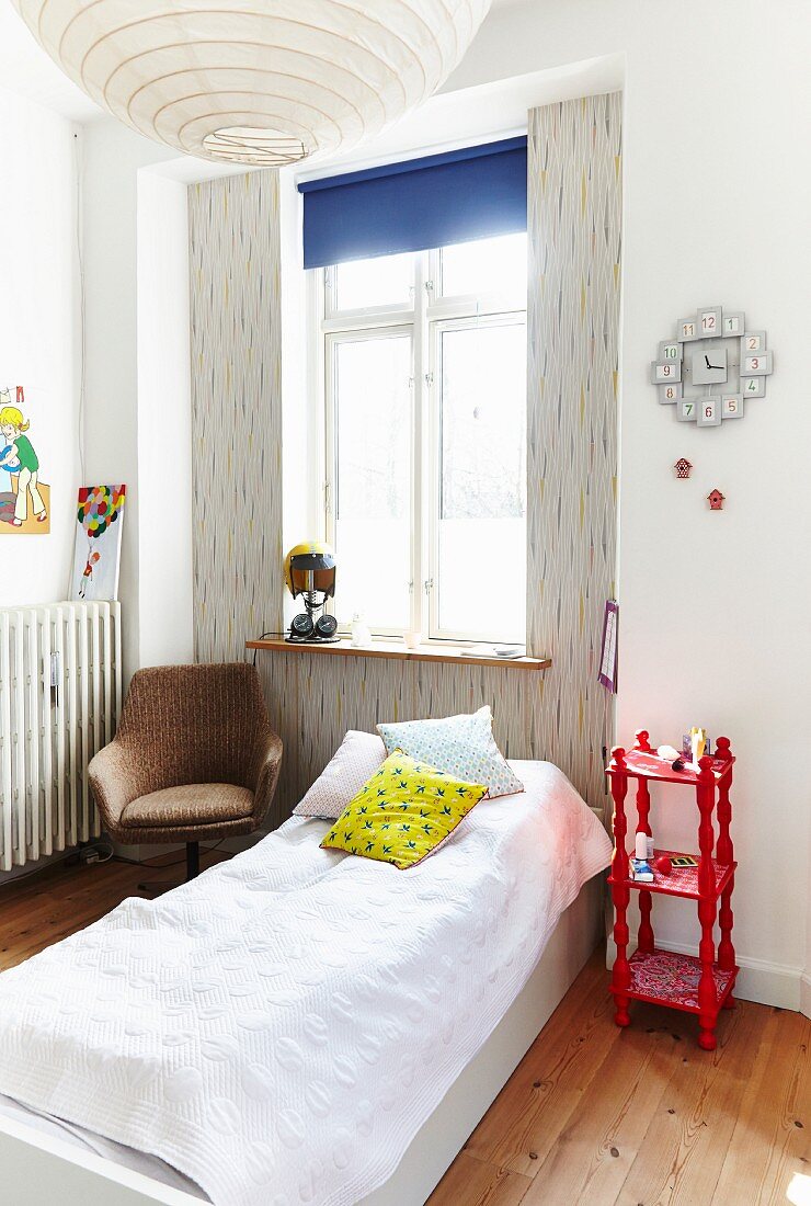 Simple bed below window, red shelving unit used as bedside table and armchair in corner