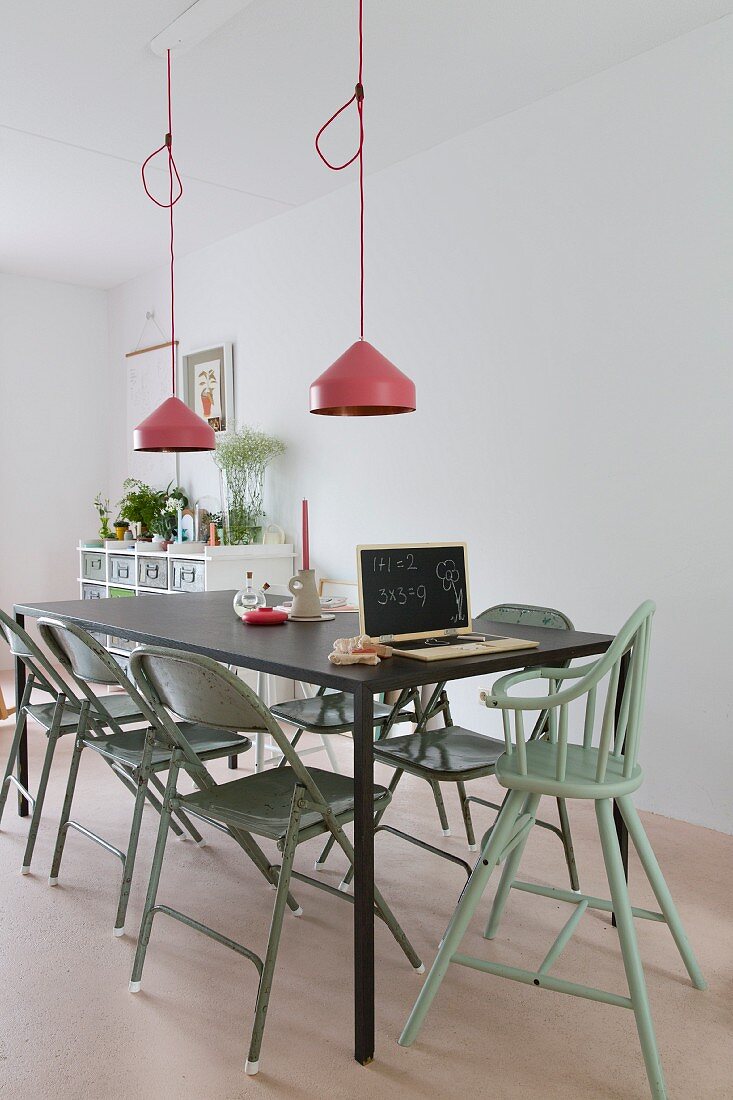 Vintage folding chairs and high chair painted green around black metal table below pendant lamps with pink lampshades