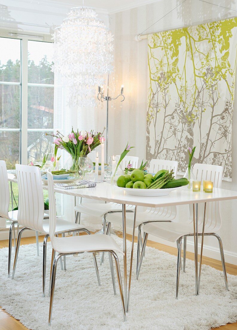 Schale mit Obst auf weißem Esstisch und moderne Stühle auf Flokatiteppich, an Wand aufgehängtes Stoffpanel