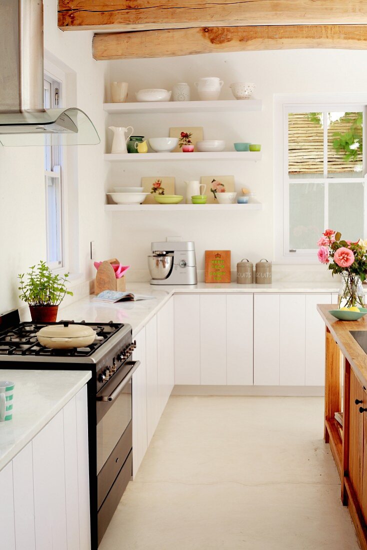 Pastellfarbenes und weißes Geschirr auf Wandregalen über weißer moderner Küchenzeile, naturbelassene Holzbalkendecke und Gasherd