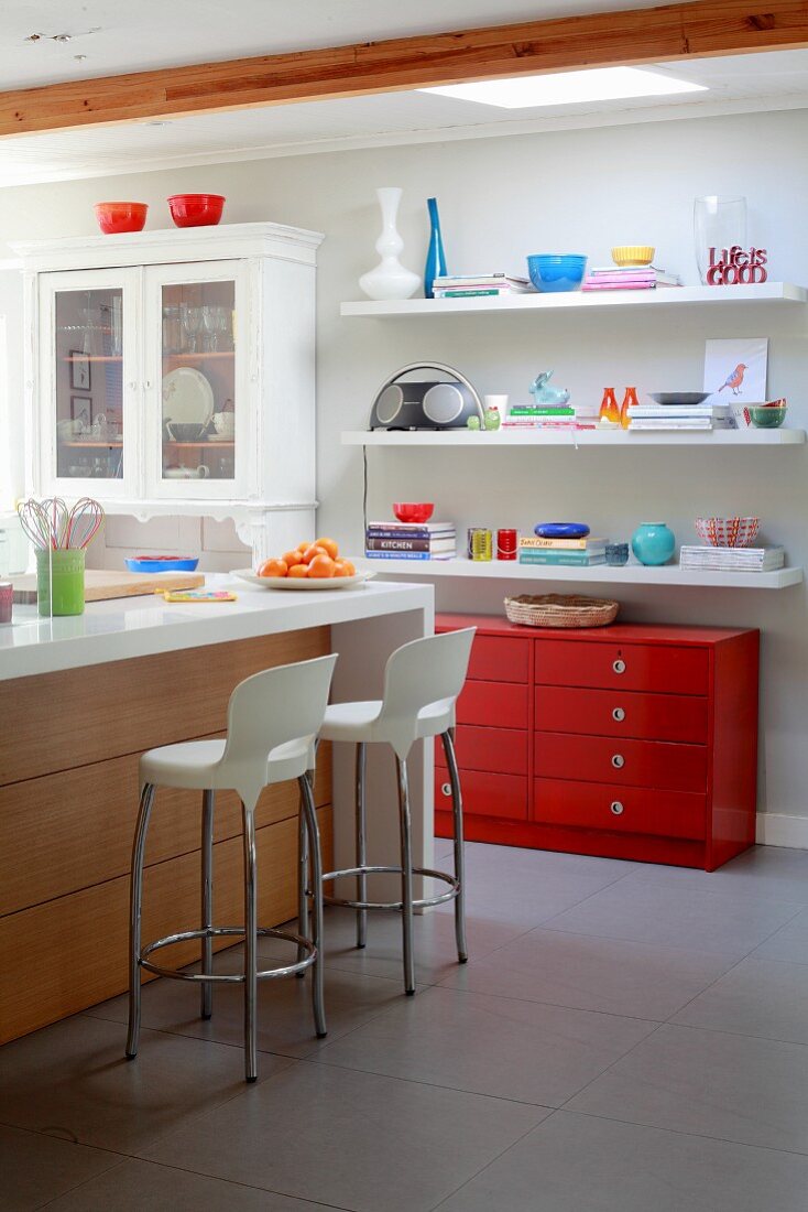 Kücheninsel und weiße Barhocker in offener Küche, an Wand gegenüber rote Schubladenkommode und weiße Regalböden mit buntem Geschirr