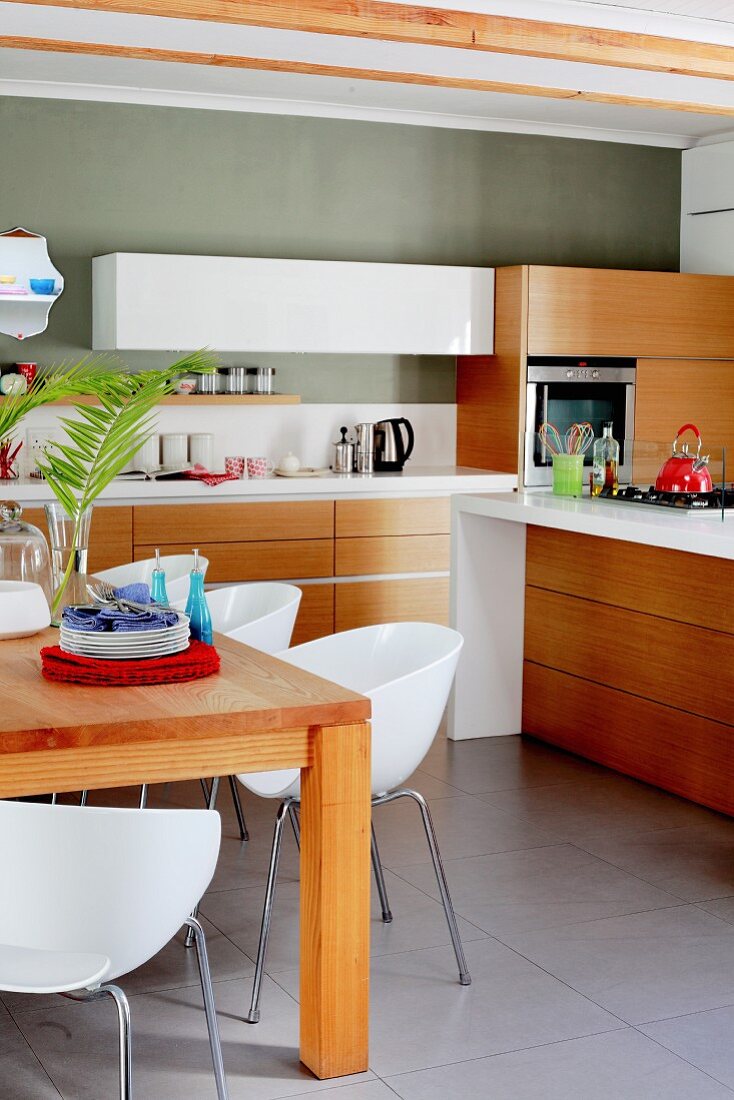 weiße Schalenstühle um Holztisch, im Hintergrund moderne Küchenzeile mit Massivholz Blenden an Unterschränken, grüngrau getönte Wand