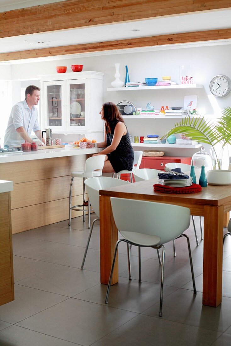 White shell chairs around wooden table and couple at modern counter with bar stools in background in kitchen with floating shelves