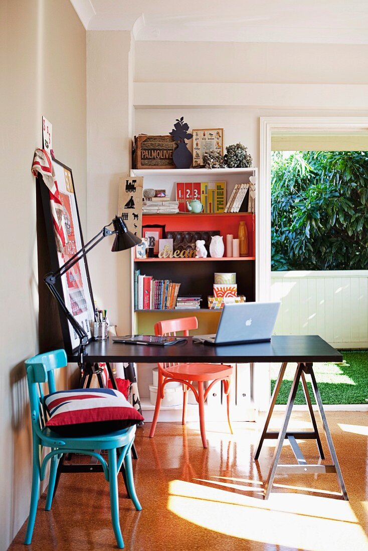Black desk on trestles, colourful wooden chairs, ornaments on bookcase and open window in background