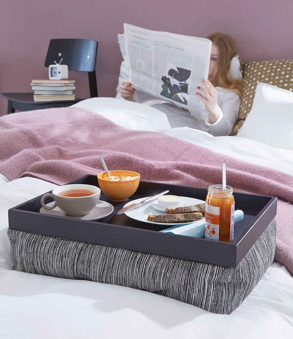 Selbst gebasteltes Kissentablett mit Frühstück im Bett, im Hintergrund Frau beim Zeitunglesen