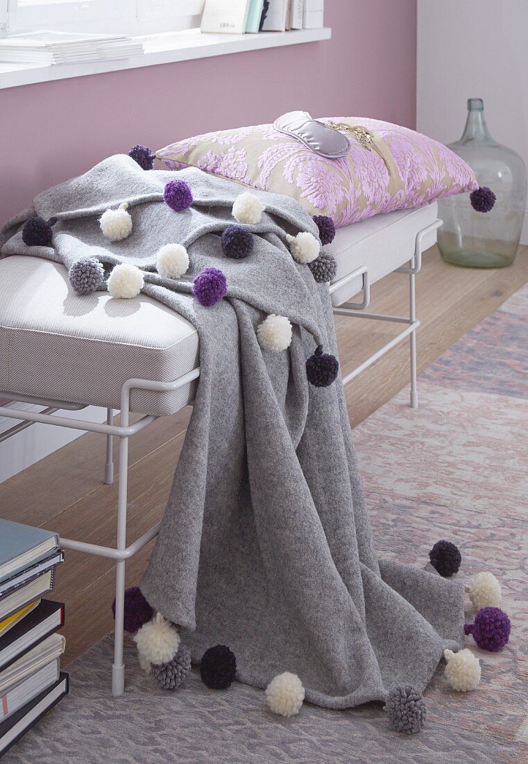 A winter throw decorated with coloured pom-poms on a day bed with a white frame against a lilac painted wall
