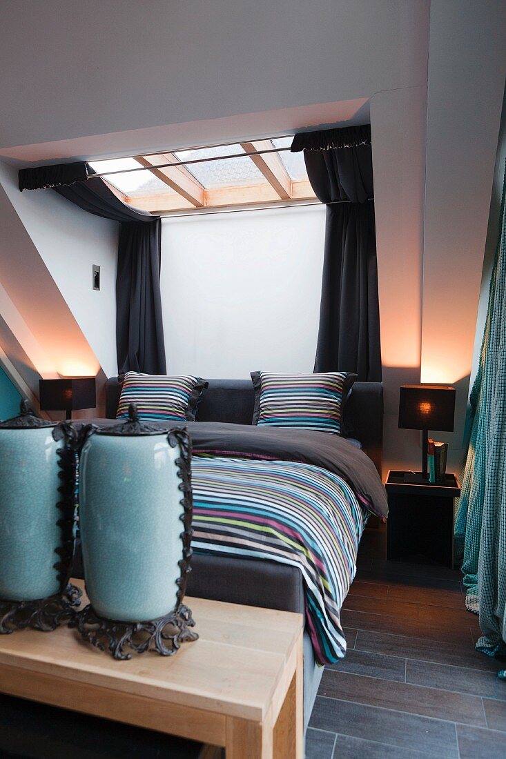 Elegantes Schlafzimmer, auf Bett Bettwäsche im Streifenmuster unter Dachfenster, im Vordergrund antike Gefässe mit Deckel