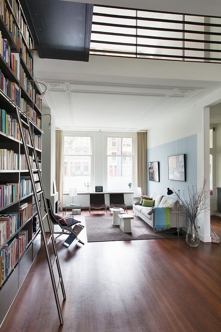 Bookcase with metal library ladder in high-ceilinged room with gallery; seating area with stucco ceiling in background