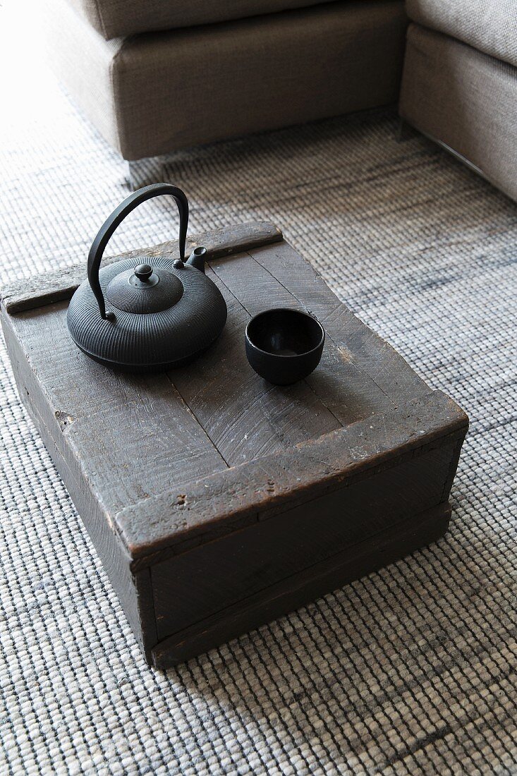 Black teapot and tea bowl on wooden crate on grey, speckled, woven carpet