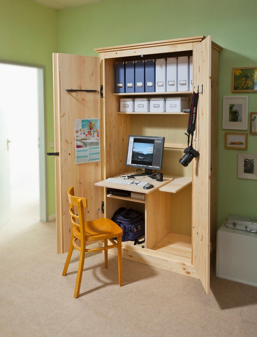 Holzstuhl vor selbstgebautem geöffnetem Büroschrank mit integrierter Schreibtischplatte, Bildschirm und Stehordnern