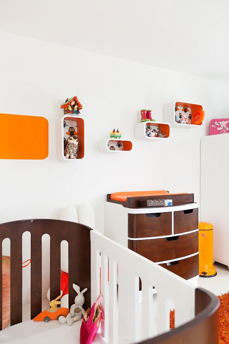 Blick über Kinderbett auf massgefertigte Kommode mit Holzschubladen, an Wand aufgehängte Regalboxen mit orangefarbenen Innenflächen