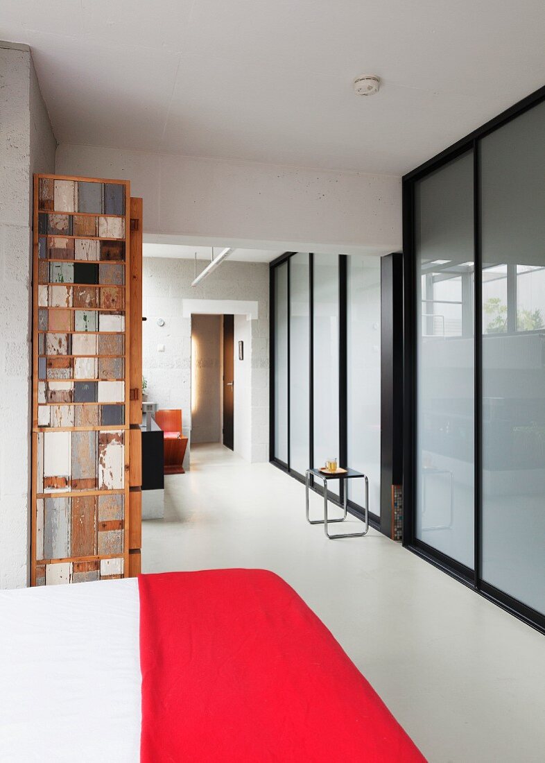 Bett mit roter Tagesdecke in offenem Wohnraum, gegenüber Wand aus satiniertem Glas und schwarzer Stahlkonstruktion