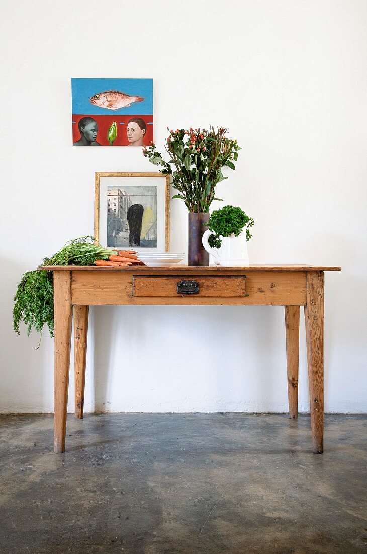 Ländlicher Holztisch mit Gemüse und Blumenvase, vor Wand mit aufgehängtem Bild