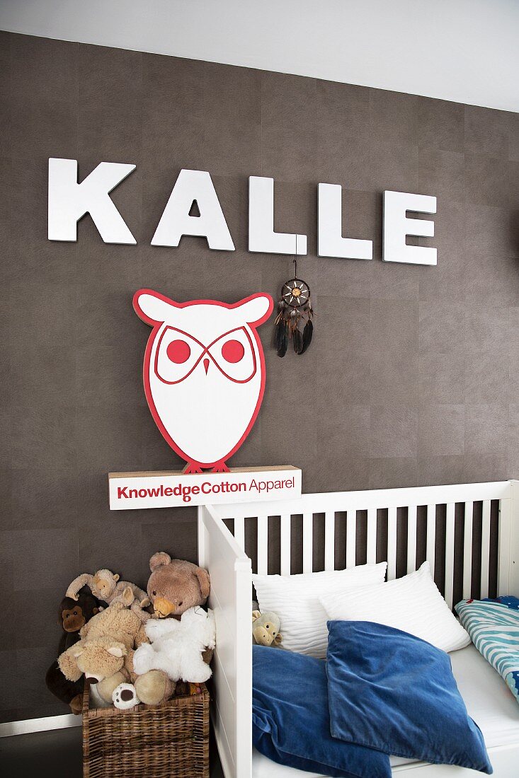 White cot with blue and white pillows below boy's name in large, white letters and owl figure on brown wall