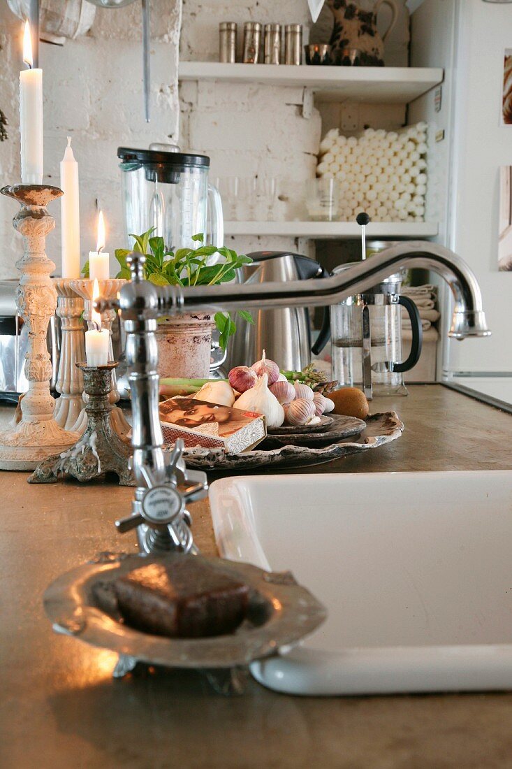 Blick auf Küchentheke mit eingebautem Spülbecken und Vintage Armatur, im Hintergrund brennende Kerzen neben Schale mit Knoblauchknollen