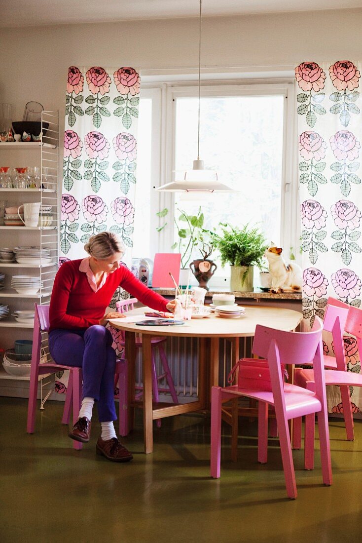 Frau auf pink lackierten Stuhl vor rundem Tisch am Fenster, bodenlange Vorhänge mit Blumenmuster