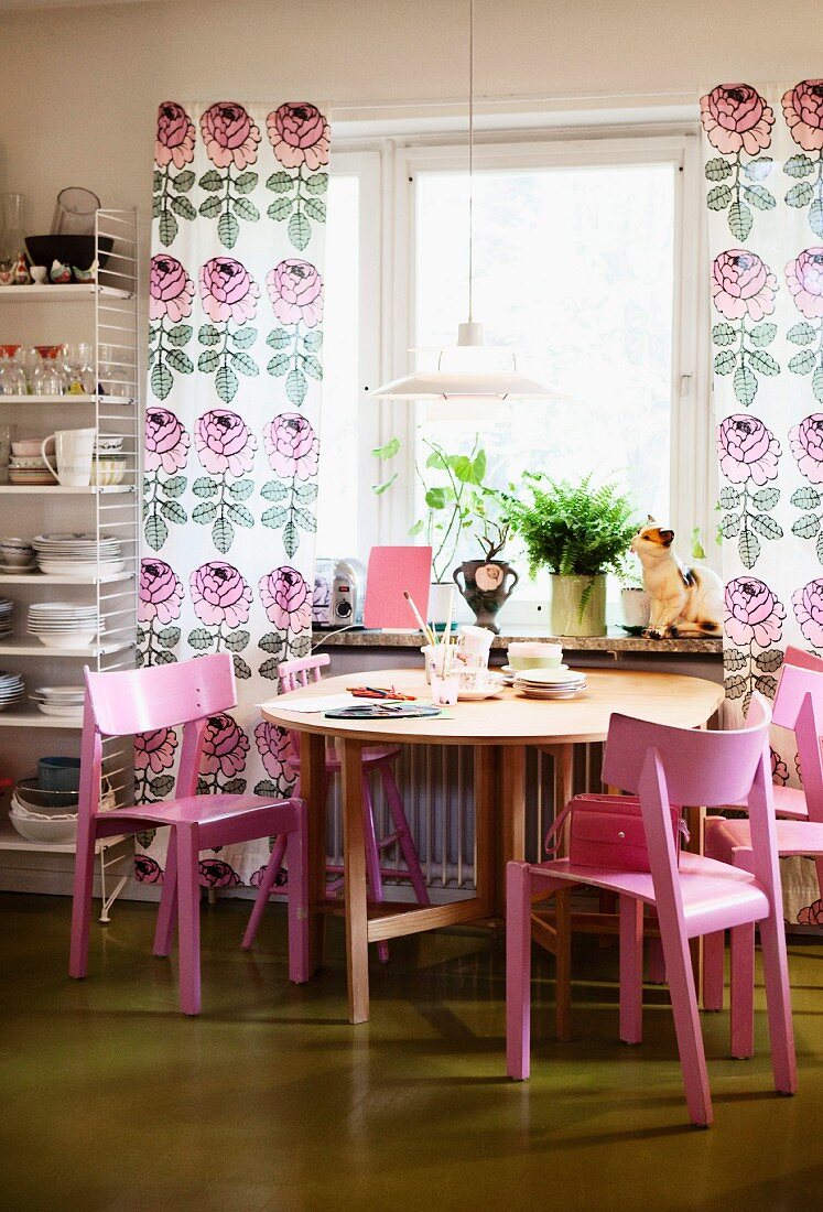 Pink wooden chairs around round table below window with floor-length, floral curtains